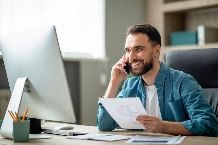 Man using his mobile phone while sitting at his desk looking at sales figures on paper.